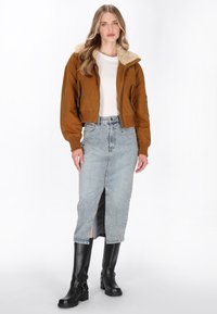 Giacca bomber marrone con colletto in pelliccia sintetica, t-shirt bianca, gonna di jeans azzurro chiaro con spacco frontale e stivali neri al ginocchio.