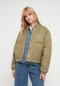 Πράσινο μπουφάν με καπιτονέ ύφασμα, κλείσιμο με κουμπιά τύπου κλιπ και δύο εμπρόσθιες τσέπες. Φορεμένο πάνω από πουκάμισο από τζιν ύφασμα, με άνετη γραμμή και ριμπ μανσέτες.