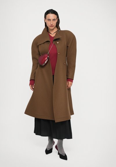 Donna che indossa un cappotto oversize marrone sopra una maglietta rossa, una gonna nera, calze grigie e tacchi neri, mentre porta una borsa a forma di cuore rossa.