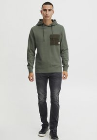 Olive green hoodie with a front pocket in dark olive, featuring drawstrings and ribbed cuffs, paired with dark jeans and black sneakers.