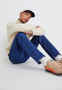 Suéter de lana beige, jeans de mezclilla azules y zapatillas color beige con acentos rojos. El modelo está sentado con los brazos alrededor de las rodillas, usando una gorra clara.
