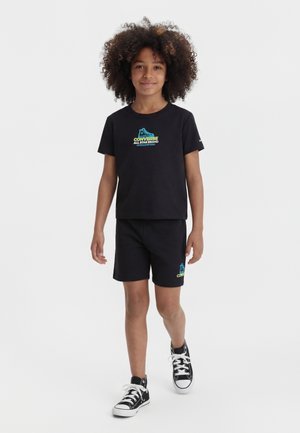 Dziecko w czarnej koszulce Converse i szortach, noszące czarne wysokie trampki, idzie do przodu na jednolitym białym tle.