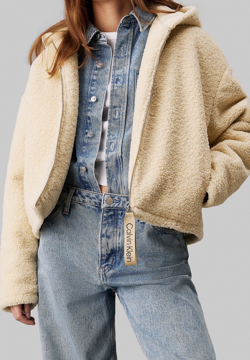 Jasno kremowy, puszysty, krótki żakiet wykonany z owczej skóry z kapturem, w połączeniu z jasnoniebieską koszulą na guziki i spodniami z wysokim stanem.