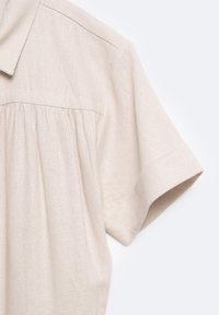Vaalean beige lyhythihainen paita, jossa on kauluspääntie, teksturoitu kangas, laskostetut yksityiskohdat etupuolella ja kääritty hihansuu.
