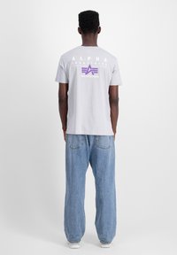 Halli puuvillane T-särk, mille seljal on suur lilla ja valge logo, koos lõdva sinise denimipükstega. Pehme tekstuur, vabalt langev lõige.