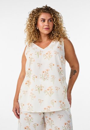 Curly-haired woman wearing sleeveless white top and pants with peach floral print, standing against plain background.