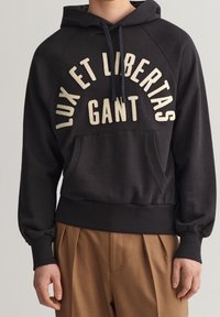 Černá mikina s kapucí a stahovací šňůrkou, se přední kapsou a krémovým textem "LUX ET LIBERTAS GANT." Hranaté manžety a lem.