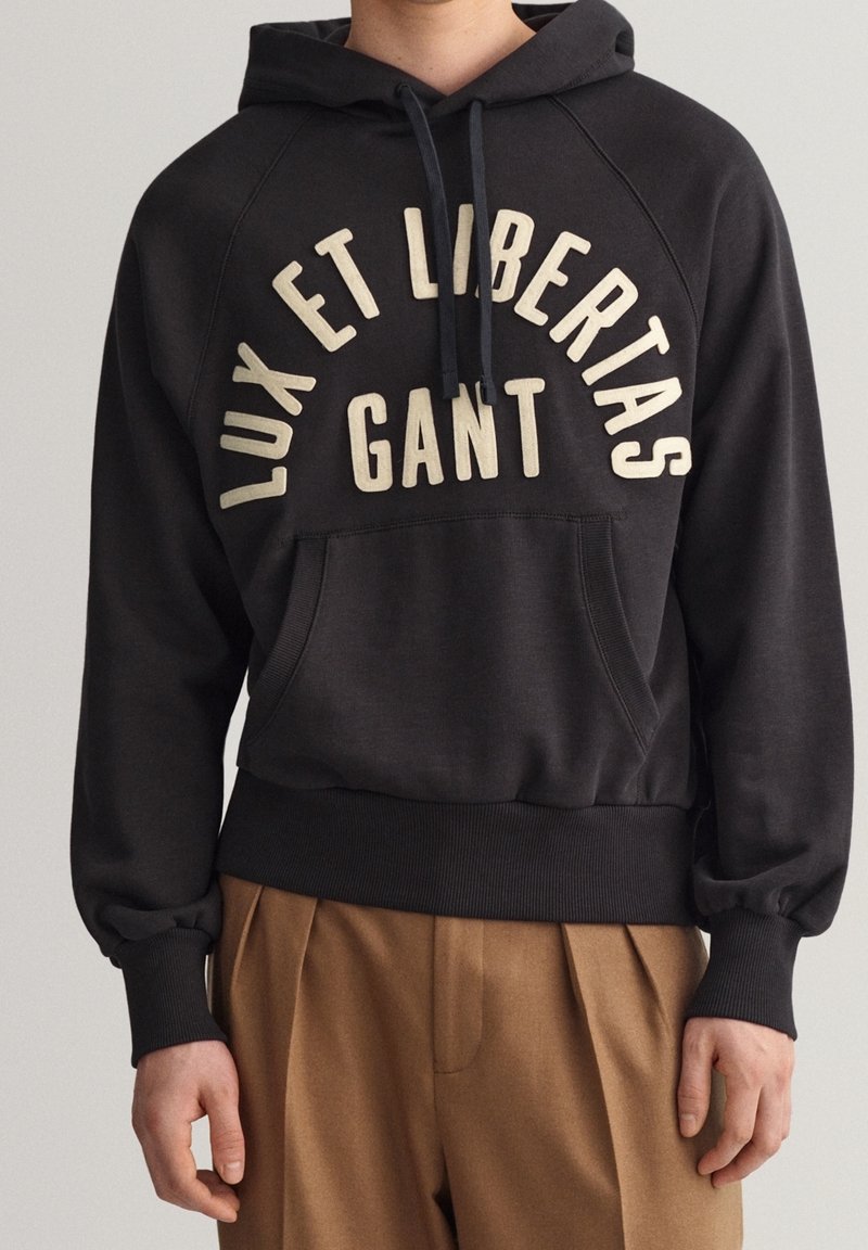 Černá mikina s kapucí a stahovací šňůrkou, se přední kapsou a krémovým textem "LUX ET LIBERTAS GANT." Hranaté manžety a lem.