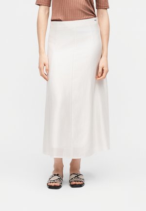 Žena stojící před jednobarevným pozadím, má na sobě bílou sukni dlouhou ke kotníkům, hnědé žebrované tričko s krátkými rukávy a černobílé pantofle s hadím vzorem.