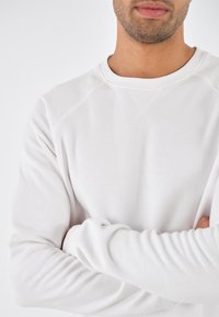 Sudadera blanca con cuello redondo, mangas largas y una textura suave. Presenta mangas raglán y detalles de costura sutiles.