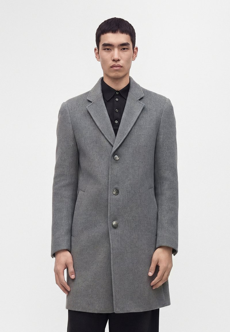 Homme portant un manteau gris à trois boutons sur une chemise noire à col, debout devant un fond clair uni.