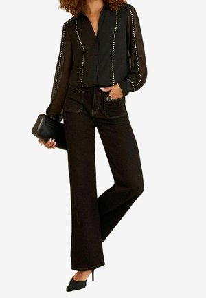 Femme portant un chemisier noir transparent clouté, jean noir à jambes larges, talons pointus noirs, tenant une pochette noire, main dans la poche.
