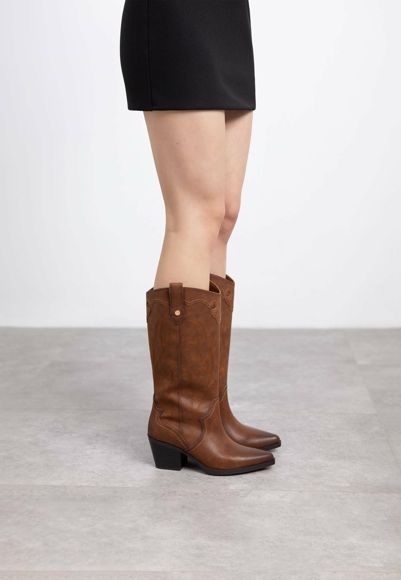 Botas de vaquero de cuero marrón con punta afilada, tacón bajo y apilado, y costuras decorativas a lo largo de los lados. Textura suave y diseño alto.