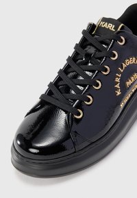 Glanz zwarte sneaker met zwarte veters, gouden veterogen en gouden "Karl Lagerfeld Paris" merknaam op de zijkant en de tong.