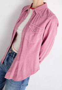 Chemise rose avec col boutonné, deux poches poitrine et boutons-pression. Confectionnée en tissu doux, elle présente une coupe décontractée et un ourlet arrondi.