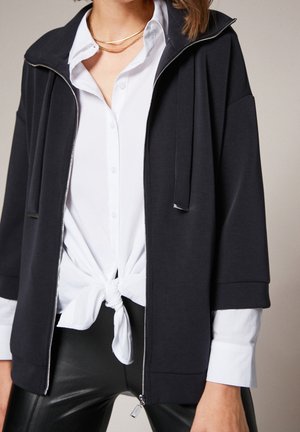 Vrouw die een zwart jack met rits draagt over een wit overhemd met knopen, gestrikt in de taille, gecombineerd met zwarte leren broek en een gouden ketting.