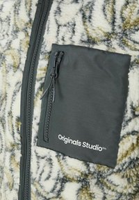 Polarowa kurtka z teksturowanym kremowo-zielonym wzorem. Posiada ciemnoszary, zapinany na zamek kieszeń na piersi z logo "Originals Studio".