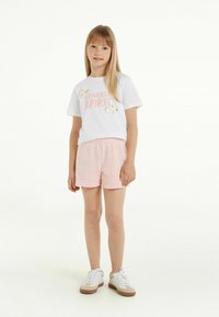 T-shirt bianca con il testo "SPARKLE SPIRIT" e grafiche a stella, abbinata a pantaloni corti rosa chiaro e sneakers bianche con dettagli beige.