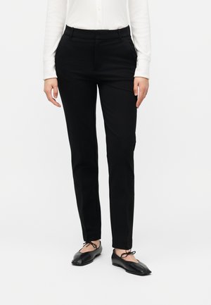 Person trägt schwarze Slim-Fit-Hose, weißes Langarmshirt und schwarze Ballettflats mit geteiltem Vorderfuß und Schnürdetails, stehend vor weißem Hintergrund.