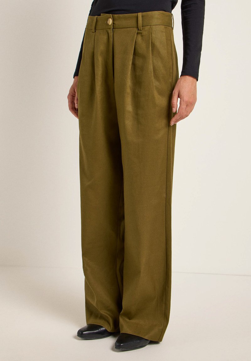 Hose mit weitem Bein aus olivgrünem Stoff, mit Falten, Knopfverschluss und glatter Textur; kombiniert mit schwarzen Schuhen.