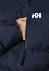 Chaqueta de plumas azul marino con cierre frontal, un bolsillo en el pecho y un logo blanco (HH) en el lado derecho. Tejido suave y ligero.