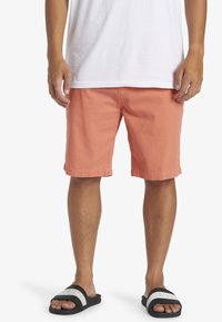 Shorts en tissu coton orange corail, avec une coupe droite, un ourlet retroussé et des poches latérales, associés à des sandales noires avec des sangles blanches.