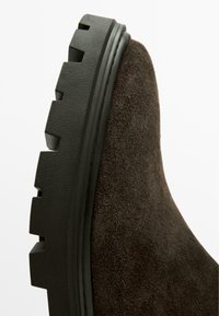 Czarny zamszowy but z teksturowaną powierzchnią, z masywną czarną gumową podeszwą o wyraźnym bieżniku, o przekroju pokazującym gładkie krawędzie.