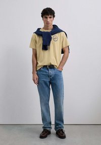 Camiseta amarilla con logo, sudadera azul marino sobre los hombros, vaqueros azules y zapatos marrones con cordones, de pie contra un fondo neutro.