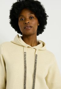 Sudadera color caqui con bolsillo frontal y acentos metálicos en los cordones. Tejido suave con un acabado texturizado y un corte relajado.