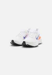 Scarpe da ginnastica bianche con tomaia in rete, swoosh viola e dettagli arancioni sul tallone, dotate di suola in gomma nera per una migliore aderenza.