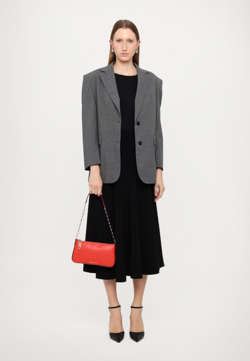 Blazer gris à rayures fines sur une robe noire, associé à un sac à main rouge et des escarpins noirs pointus. La tenue présente des lignes structurées et des textures lisses.