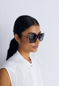 Ochelari de soare tip cat-eye cu lentile întunecate, accente metalice și un design elegant. Purtați cu o bluză albă cu guler, cu pliuri.
