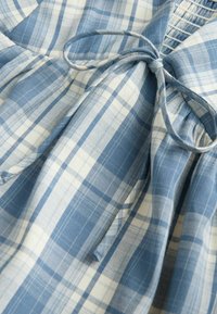 Tissu à carreaux bleu clair et blanc avec un détail de nœud noué, montrant la texture et les plis superposés.