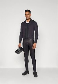 Čierny cyklistický outfit pozostávajúci z dlhého dresu s dlhými rukávmi a priliehavých legín. Vpredu má textúrovaný panel a drží čiernu prilbu.