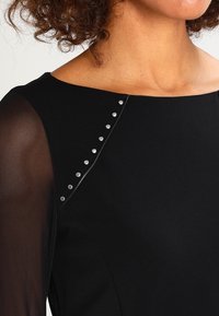 Schwarzes Kleid mit transparenten langen Ärmeln, strukturiertem Ausschnitt und silbernen Nieten entlang der Schulternaht für einen zusätzlichen Akzent.