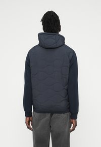 Μπλε ναυτικό μπουφάν με επένδυση και υφή με κυματιστό σχέδιο, με κουκούλα και μακριά μανίκια, συνδυασμένο με σκούρα γκρι τζιν.