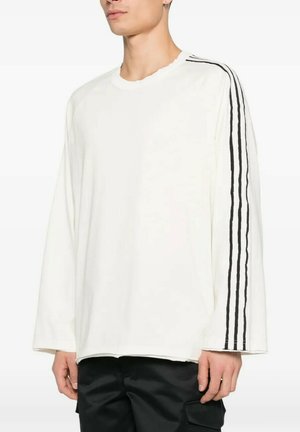 Persona che indossa una camicia bianca a maniche lunghe e larga con tre strisce nere che corrono dalla spalla al polso sul braccio destro e pantaloni neri.