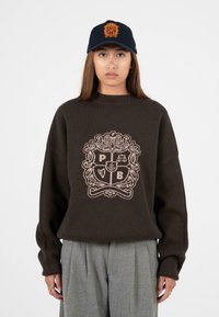 Hnědý pletený svetr s vysokým výstřihem a oversized střihem, zdobený jemným béžovým logem. V kombinaci se šedými skládacími kalhotami a námořnickou čepicí.