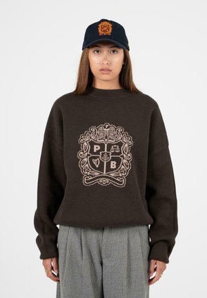 Maglione a maglia marrone con un alto collo e una vestibilità oversize, caratterizzato da un design del logo beige chiaro. Abbinato a pantaloni grigi plissettati e un berretto blu navy.