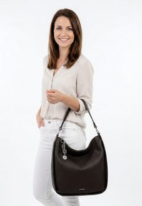 Šypsosi moteris, vilkinti lengvą palaidinę ir baltas kelnes, laiko didelę juodą tekstūruotą peties rankinę su sidabriniais pakabukais ant balto fono.