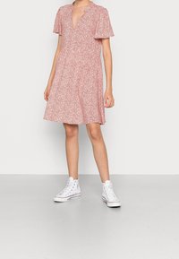 Rotes Blumenkleid mit kurzen, schwingenden Ärmeln und V-Ausschnitt. Die A-Linien-Form endet oberhalb des Knies. Kombiniert mit weißen High-Top-Sneakern.