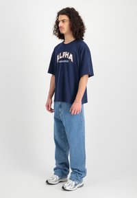 Navyblauwe T-shirt met korte mouwen met de grafische tekst "ALPHA INDUSTRIES", gecombineerd met lichtblauwe baggy jeans en zilveren sportschoenen.