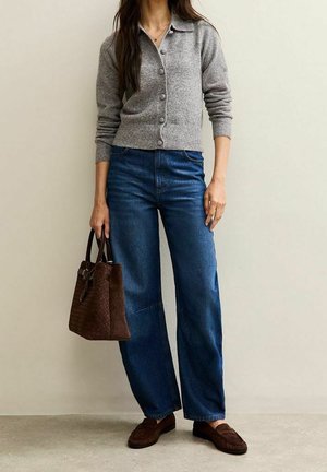 Femme portant un cardigan gris boutonné et un jean bleu à jambes larges, tenant un sac à main marron et portant des mocassins marron, debout contre un mur uni.