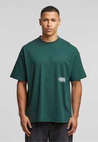 Grønn bomull t-skjorte med korte ermer, rund hals, og en liten hvit grafisk logo på venstre side. Avslappet passform og ensfarget.