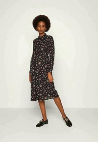 Vestido preto floral com mangas longas, frente com botões e detalhe na cintura. Combinado com mocassins pretos para um look completo.