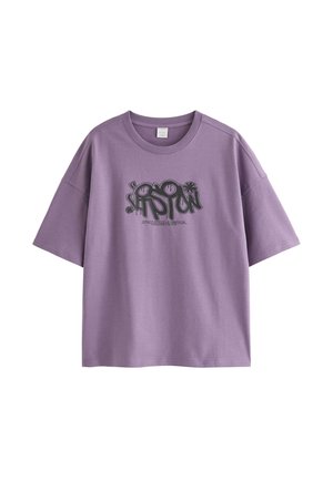 Ung person med blondt hår iført en lilla grafisk t-shirt, grå jeans, beige sneakers og en sølvkæde, der står mod en hvid baggrund.