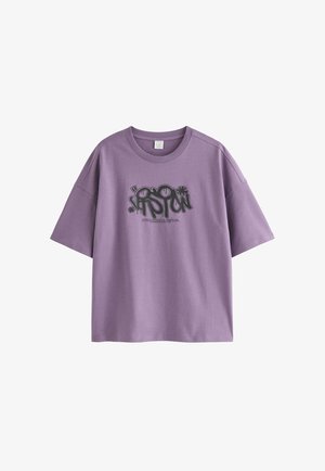 Ung person med blondt hår iført en lilla grafisk t-shirt, grå jeans, beige sneakers og en sølvkæde, der står mod en hvid baggrund.