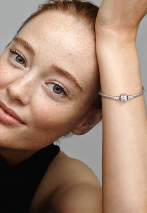 Primo piano di una donna sorridente con capelli rossi e lentiggini che indossa un braccialetto d'argento con un ciondolo sul polso sollevato.