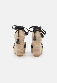 Sandales compensées espadrilles avec une tige en tissu noir, un talon en jute naturel et des rubans noirs avec des franges à l'arrière. Détail à motif à carreaux.