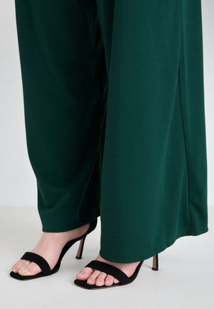 Benen in donkergroene wijde pantalons met zwarte hoge hakken met een enkele band over de tenen op een lichte achtergrond.